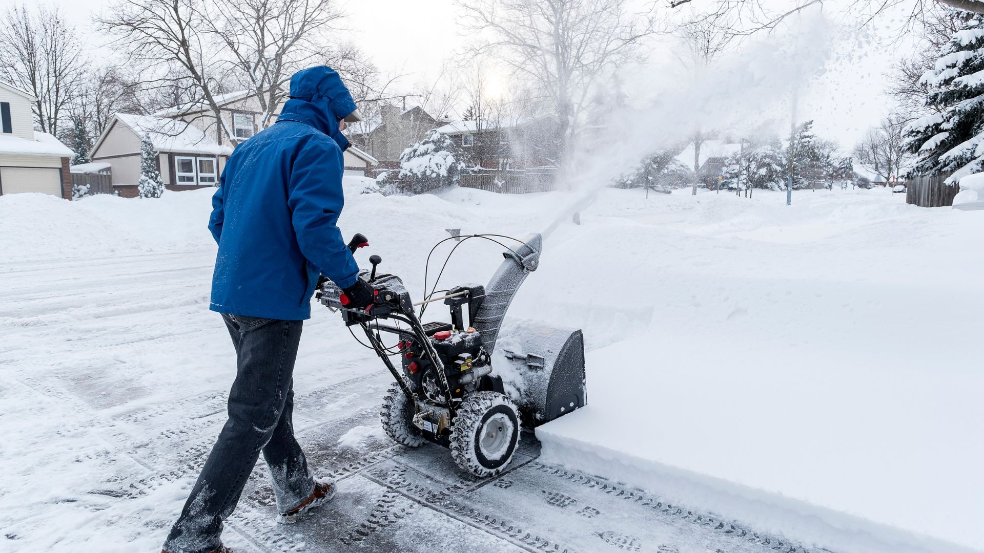 Person in blue jacket using snow blower to clear snowy suburban street